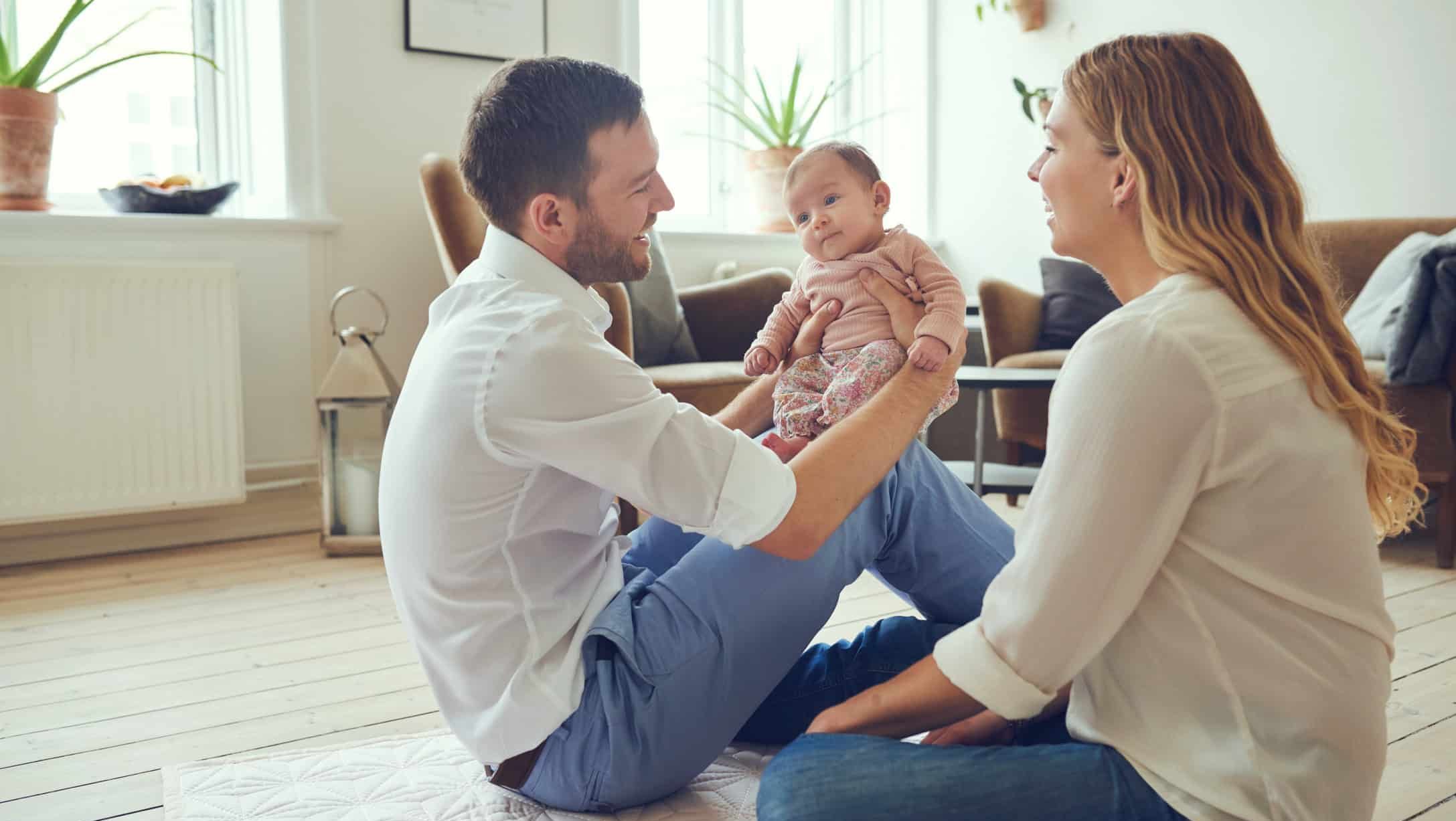 Voksen holder spedbarn på lekematte i stue, mens annen voksen sitter ved siden av i hjemmemiljø.